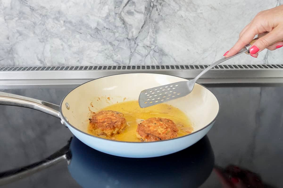 Two Salmon patties in fry pan with oil, hand holding a metal spatula.