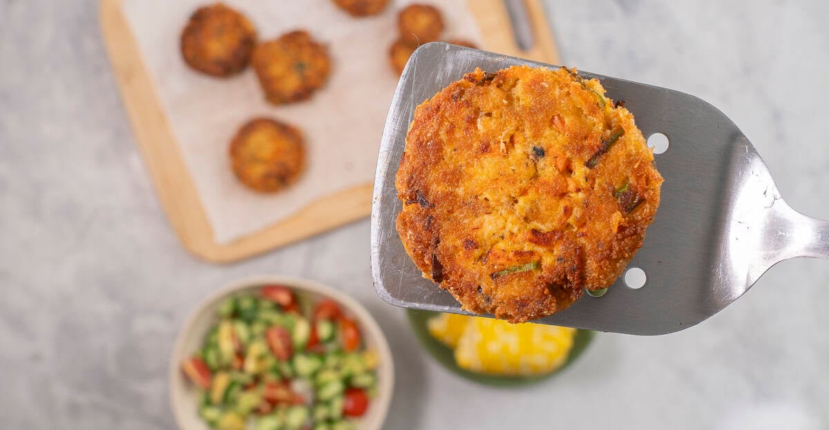 Metal spatula with salmon pattie held up, wooden chopping board with remainder of patties, bowl of fresh salad and corn below.