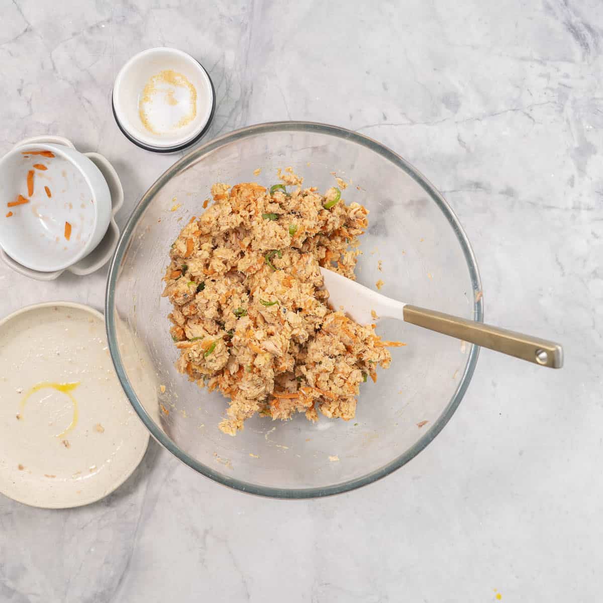 Pattie mixture in big glass bowl with spatula inside, empty ramekins from ingredients on bench top.