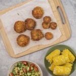 Wooden chopping board with salmon patties, bowl with fresh salad and corn on the cobs.