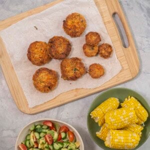 Wooden chopping board with salmon patties, bowl with fresh salad and corn on the cobs.