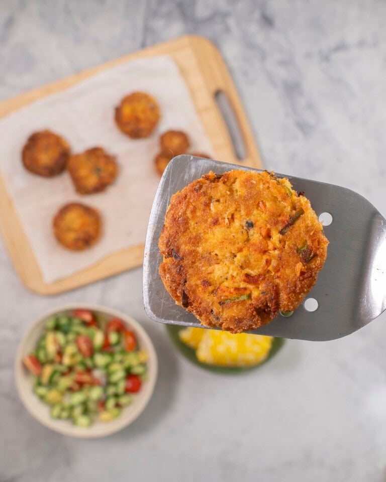 Metal spatula with salmon pattie held up, wooden chopping board with remainder of patties, bowl of fresh salad and corn below.