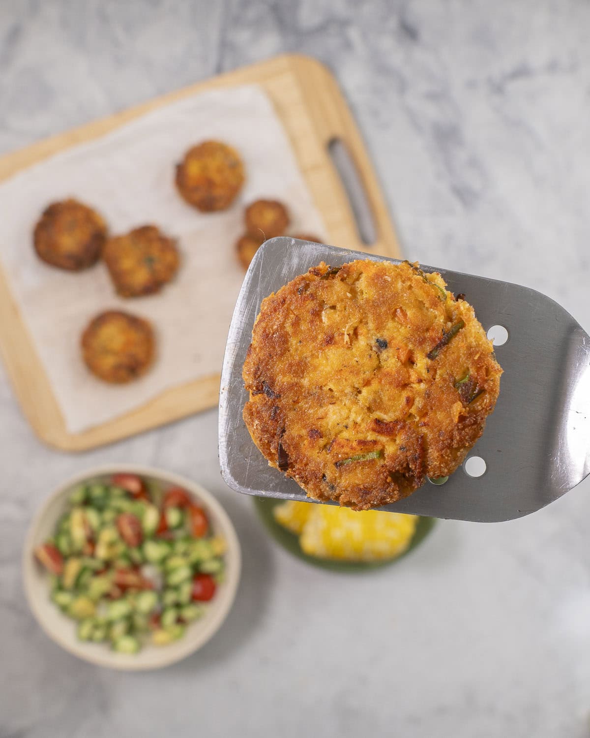 Metal spatula with salmon pattie held up, wooden chopping board with remainder of patties, bowl of fresh salad and corn below.