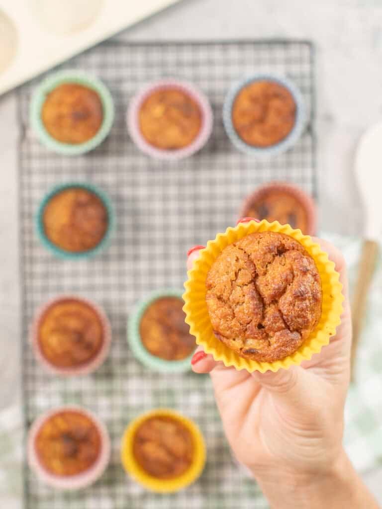 Hand holding a weebix muffin in yellow silicone mold above cooling tray with rest of muffins below.