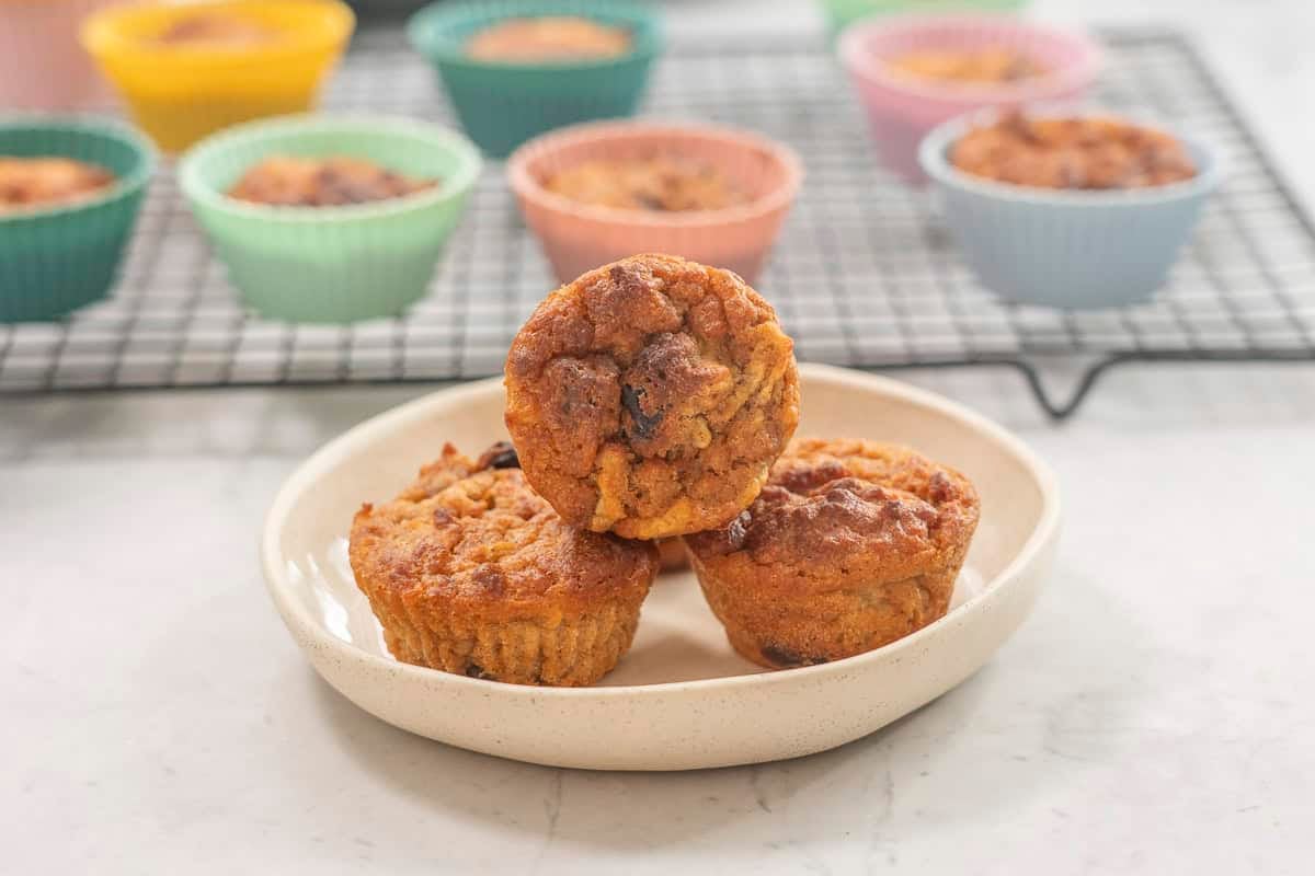 Three weetbix muffins on a plate, cooling tray with muffins in background.