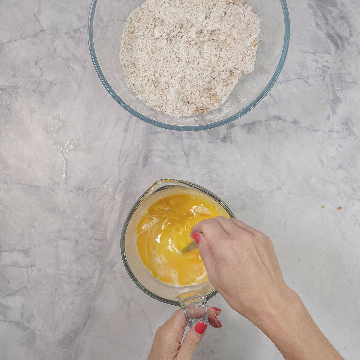 Large glass bowl of dry ingredients and small jug with hand whisking egg together with fork on benchtop.