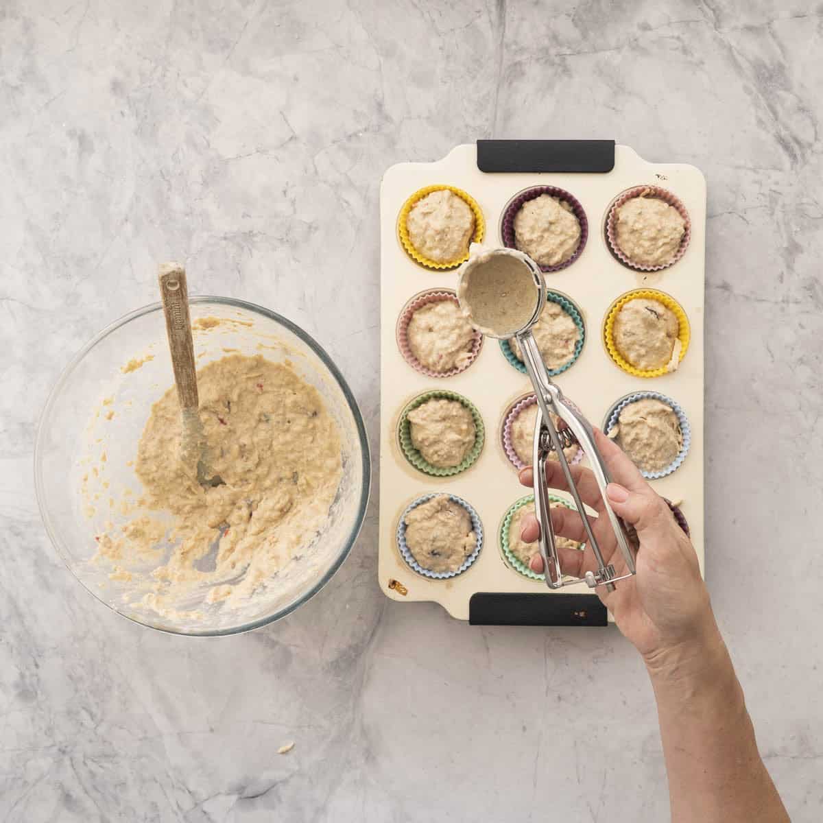 Hand holding a cookie scoop with weetbix mixture above a muffin tray filled with mixture, big glass bowl with remainder mixture inside.