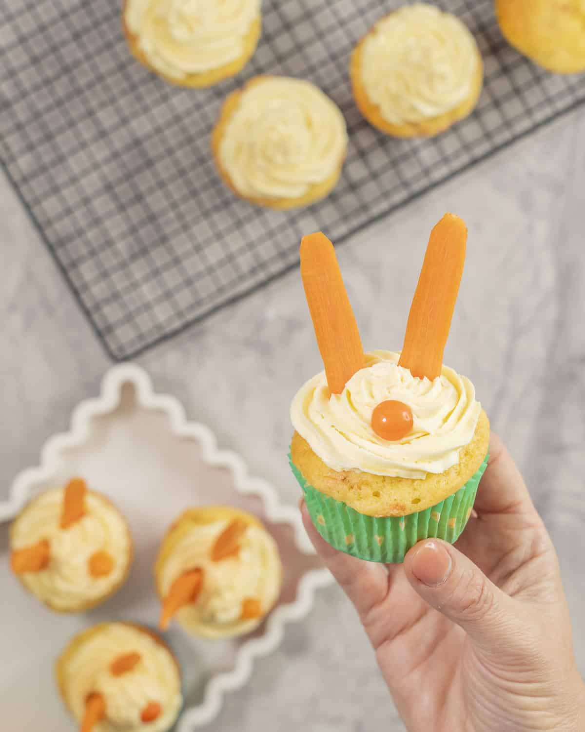Woman's hand holding a cupcake with icing piped on top, carrot sticks as ears and an orange lolly as a nose.