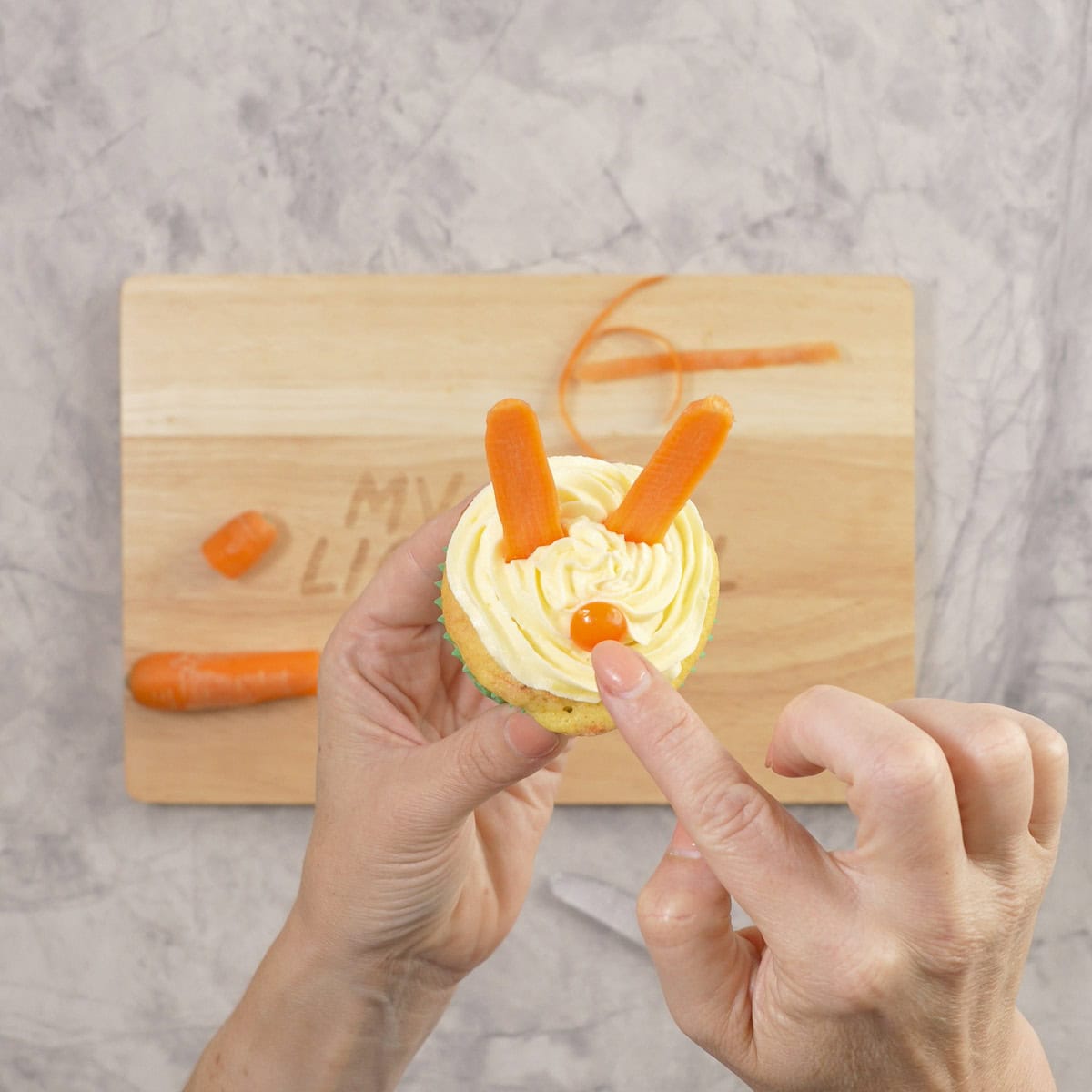 Womans hand holding a carrot cupcake, placing a orange lolly onto icing as a nose.