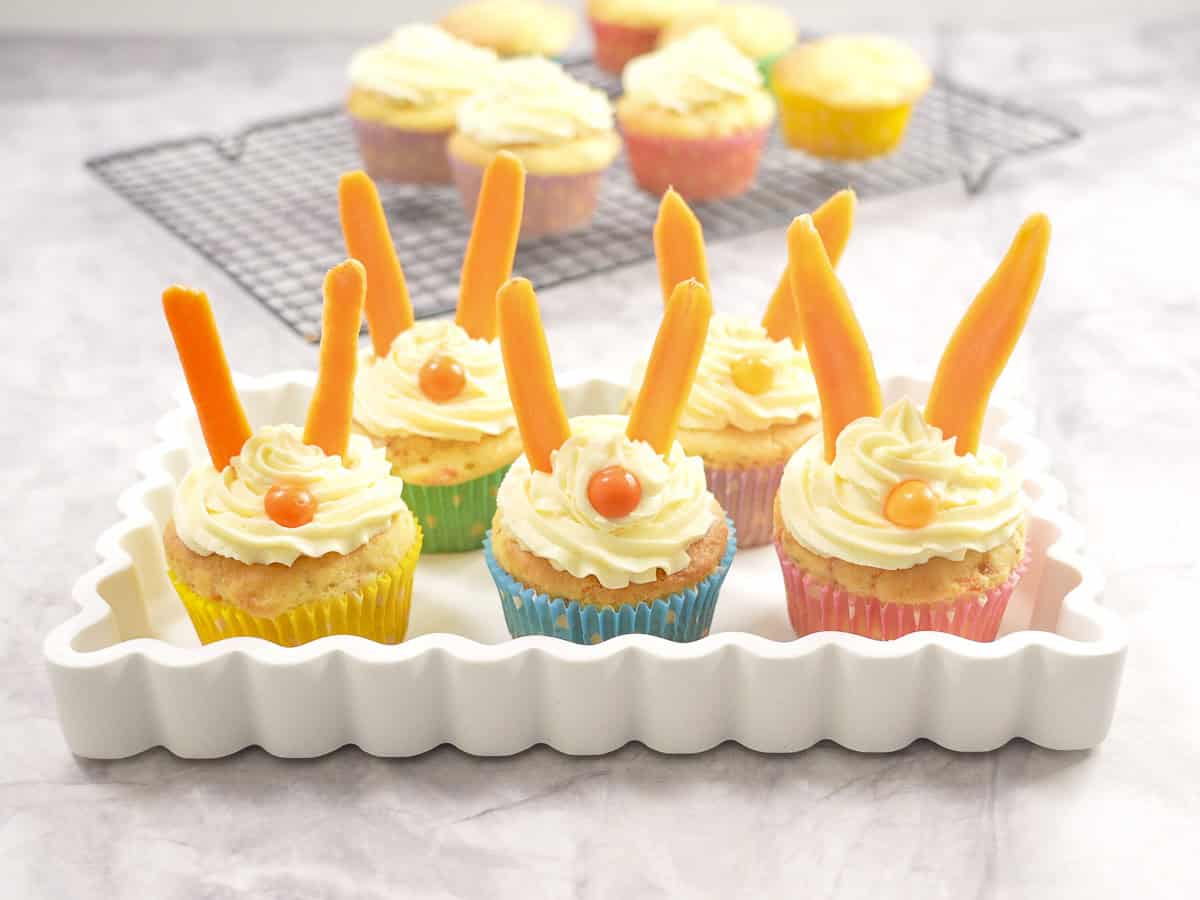 Five easter bunny carrot cupcakes with piped icing, carrot sticks as ears and orange lollies as noses on a curved serving tray.