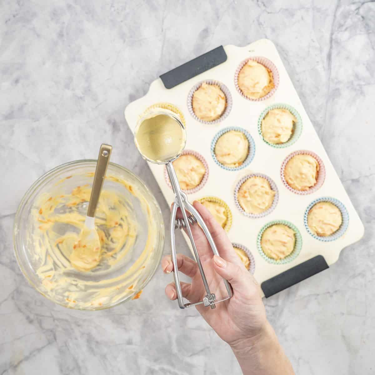 Woman holding s cookie scooper, scooping batter into a muffin tray lined with colourful cupcake liners.