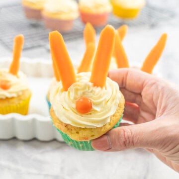 Woman's hand holding a cupcake with icing piped on top, carrot sticks as ears and an orange lolly as a nose.