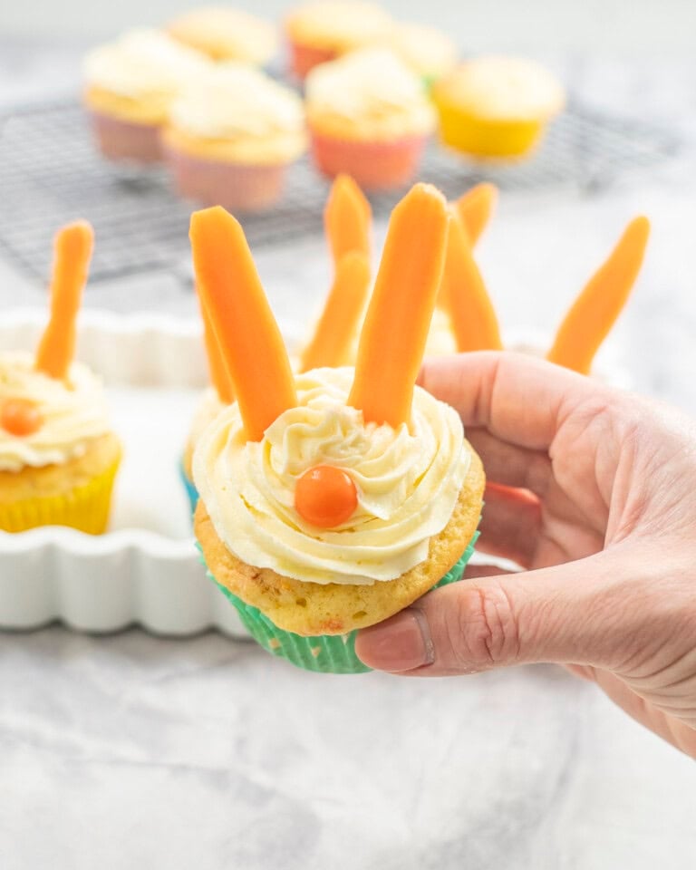 Woman's hand holding a cupcake with icing piped on top, carrot sticks as ears and an orange lolly as a nose.