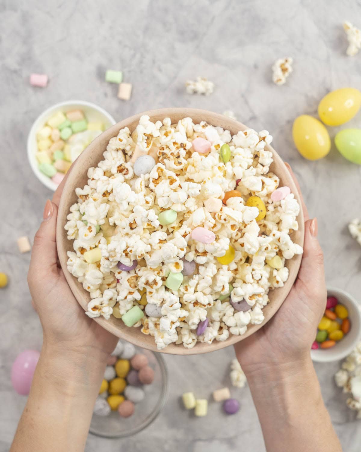 Womans hands holding a large bowl of popcorn with lollies scattered throughout.