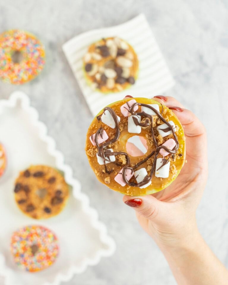 Womans hand holding a green apple with core removed sliced to look like a donut, spread with peanut butter and topped with cookie crumb, mini pink and white marshmallows and drizzled melted chocolate. 5 other apple donuts below with various toppings.