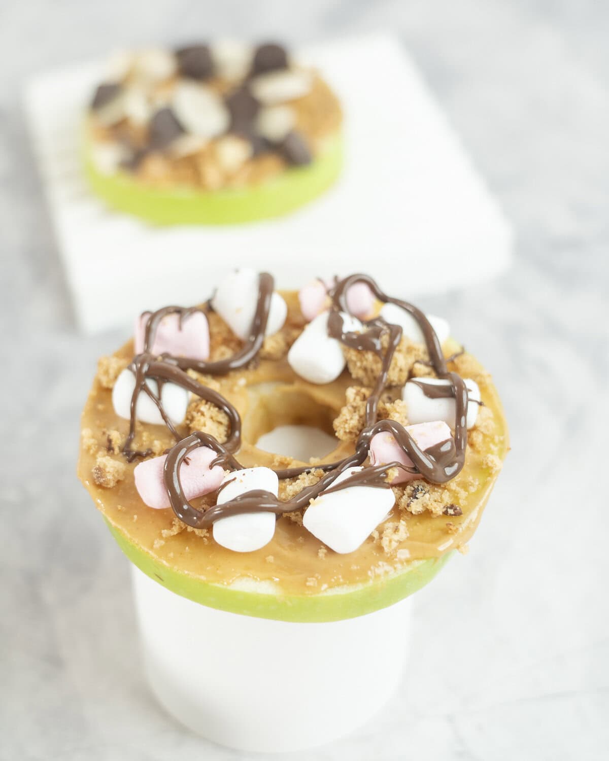 A green apple with core removed cut in a donut shape, spread with peanut butter, topped with cookie crumb, mini pink and white marshmallow and drizzle of chocolate.