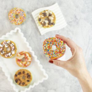 Womans hand holding a red apple with core removed sliced to look like a donut, spread with peanut butter and topped with sprinkles. 5 other apple donuts below with various toppings.