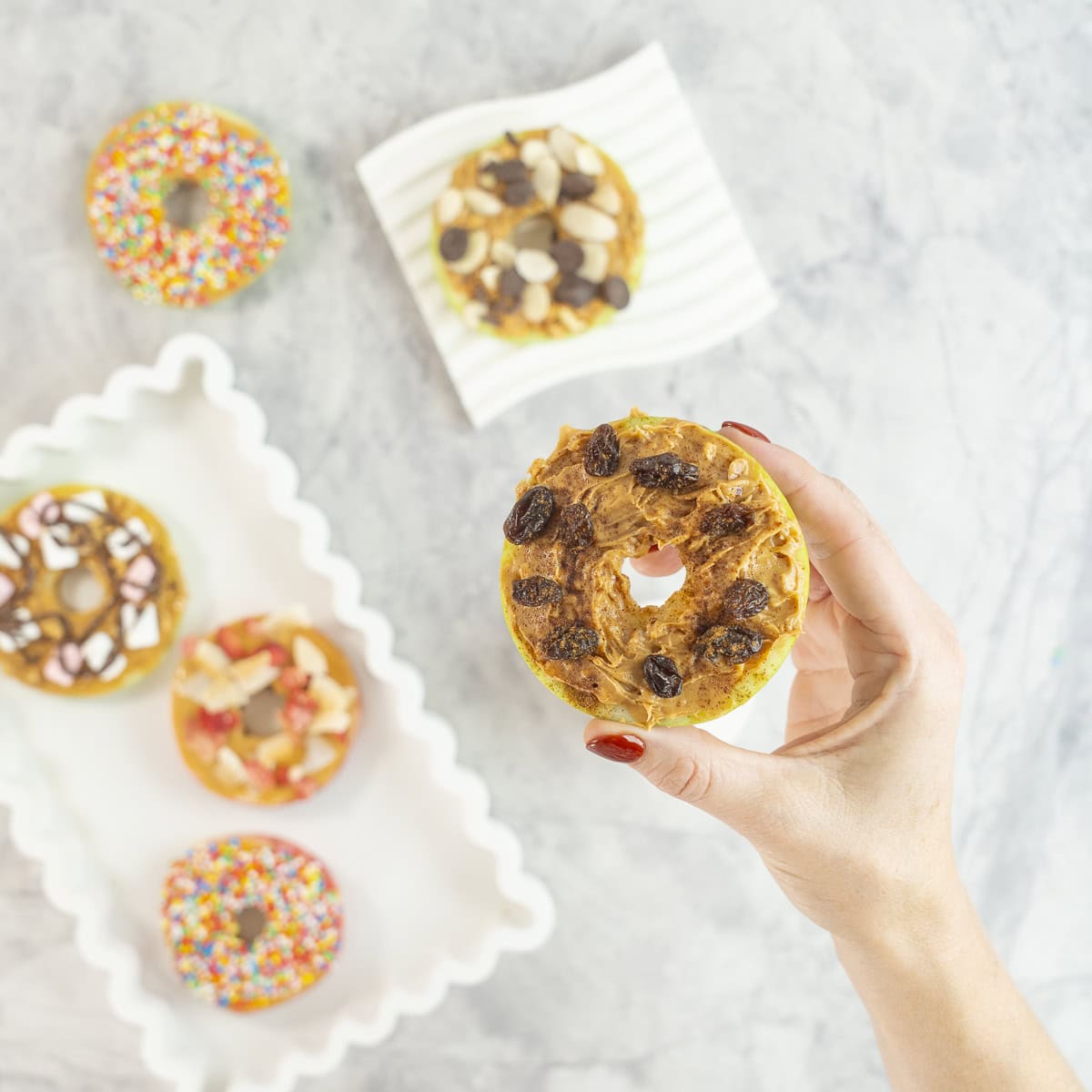 Womans hand holding a green apple with core removed sliced to look like a donut, spread with peanut butter and topped with raisins. 5 other apple donuts below with various toppings.
