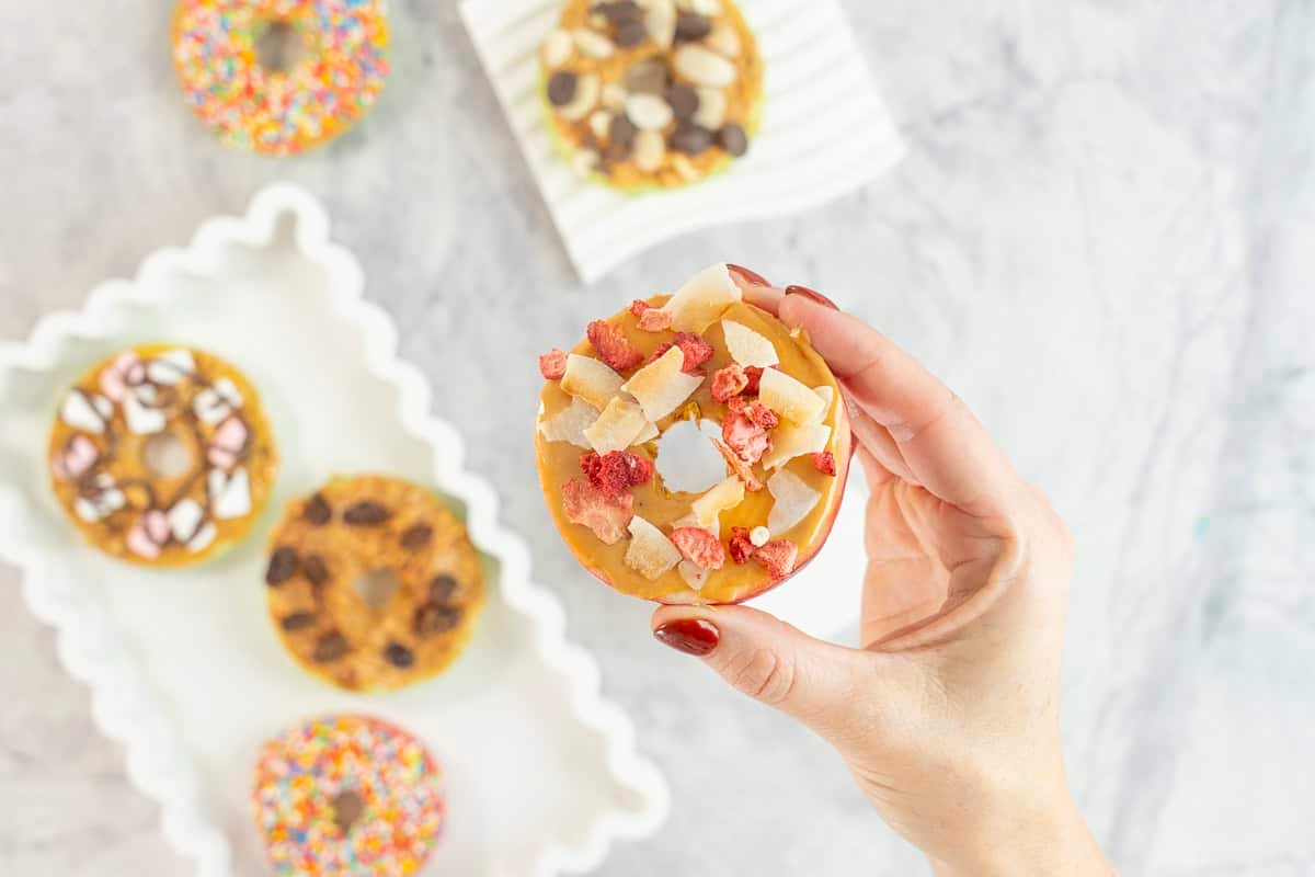 Womans hand holding a red apple with core removed sliced to look like a donut, spread with peanut butter and topped with dried raspberries coconut flakes.. 5 other apple donuts below with various toppings.