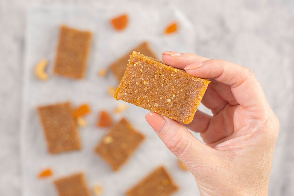 Womans hand holding a piece of apricot bar, baking paper on benchtop below with rest of apricot bars.