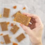 Womans hand holding a piece of apricot bar, baking paper on benchtop below with rest of apricot bars.