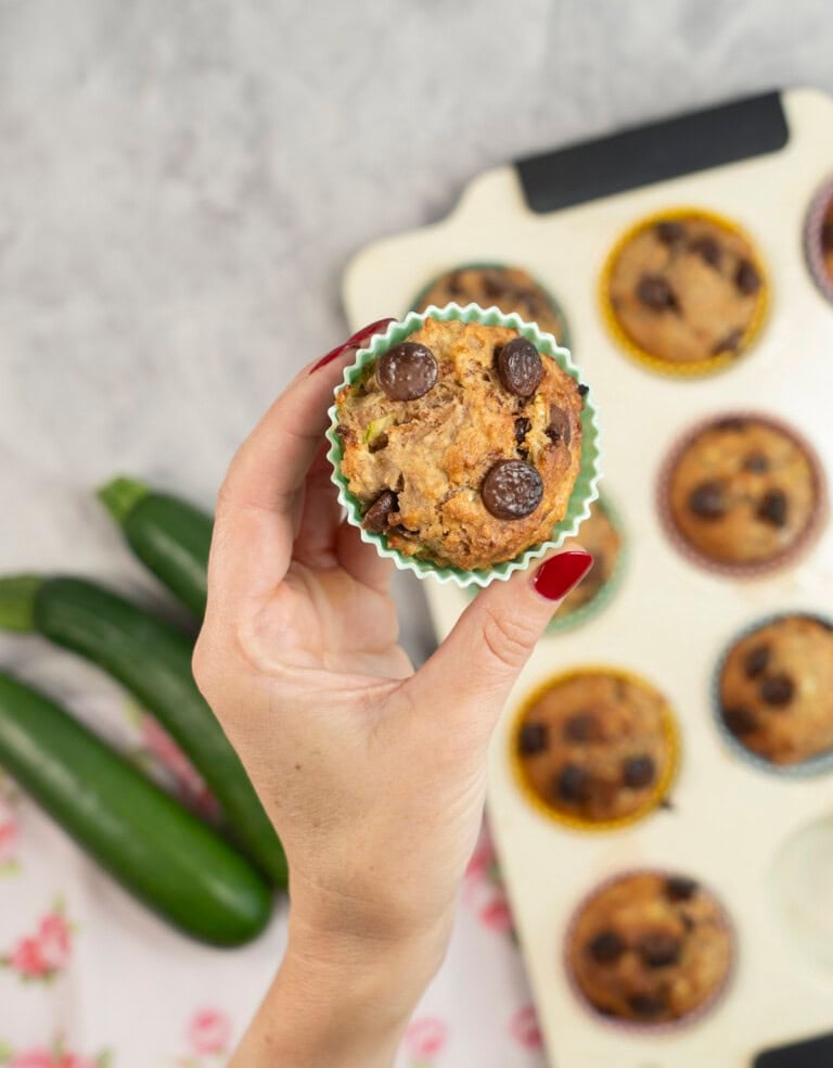 Womans hand holding a Zucchini Muffin up above a baking tray with others below on benchtop.