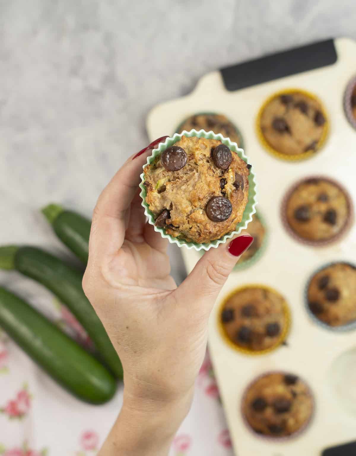 Womans hand holding a Zucchini Muffin up above a baking tray with others below on benchtop.