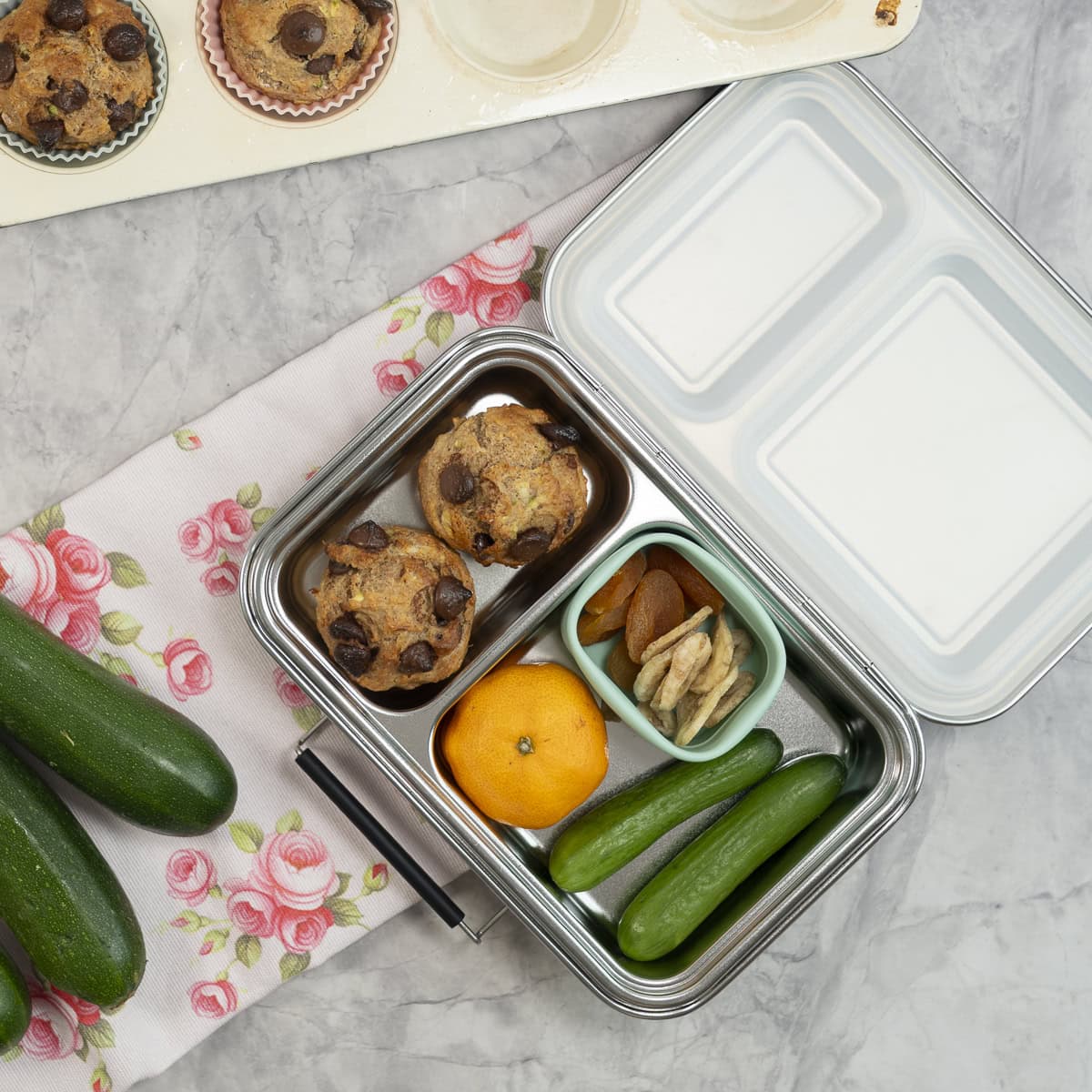 Stainless steel lunch box open on benchtop with two banana zucchini muffins inside, mandarin, mini cucumber, dried apricot and banana.