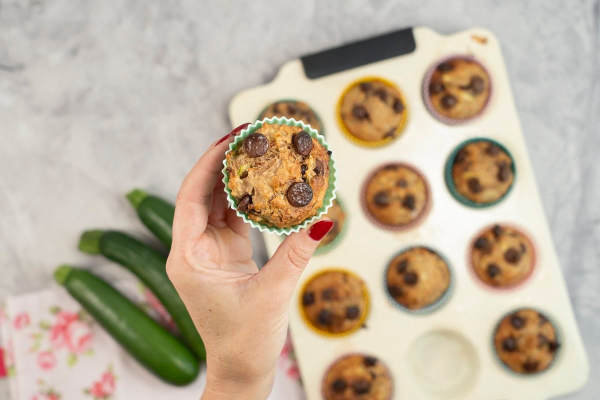 Womans hand holding a Zucchini Muffin up above a baking tray with others below on benchtop.