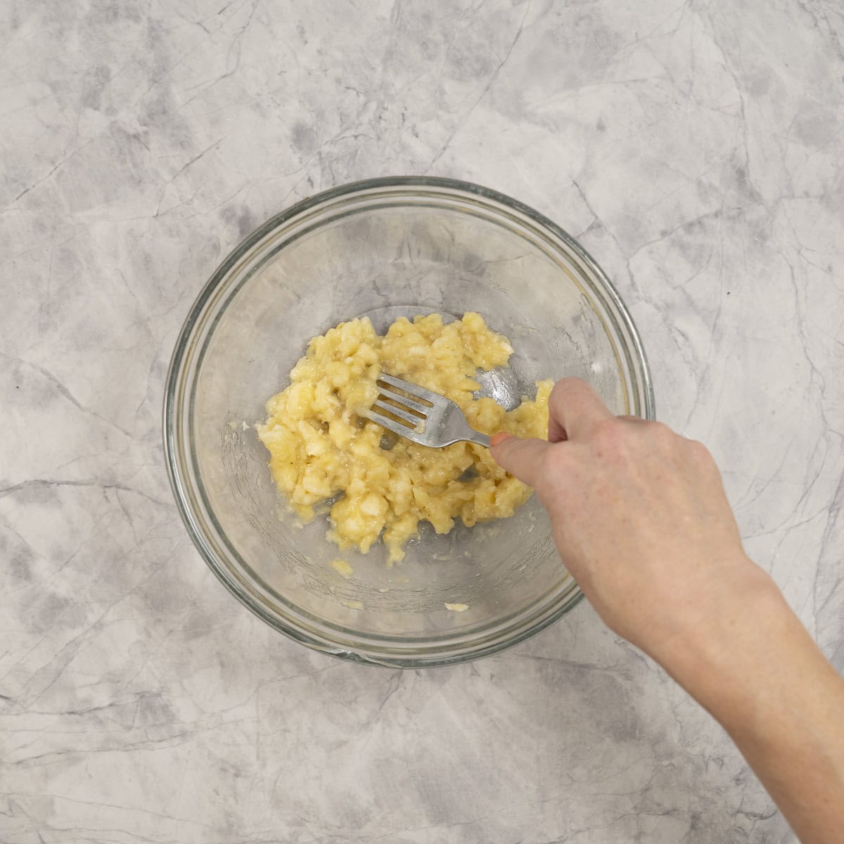 Hand holding a fork mashing banana in a glass bowl.