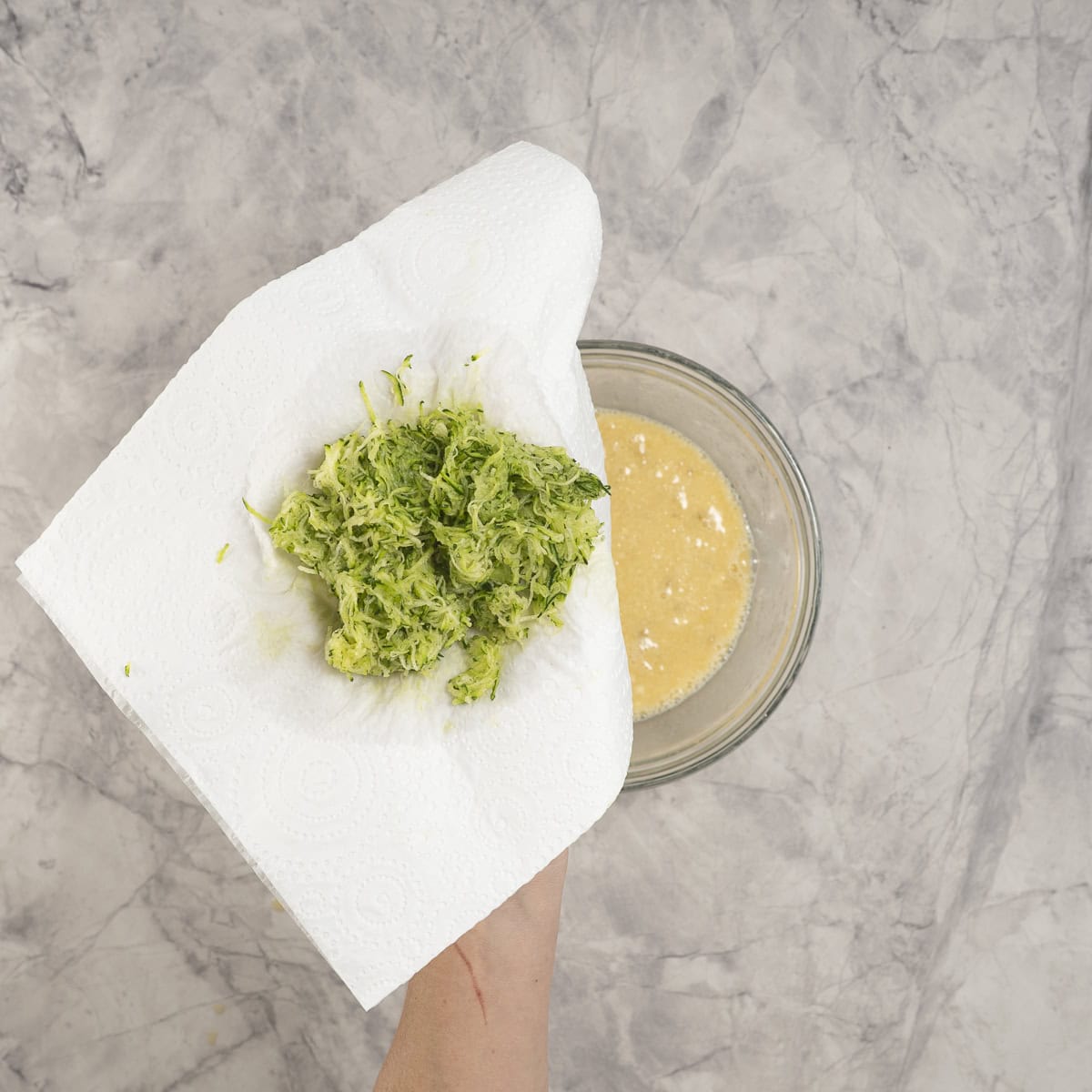 Hand holding grated zucchini in a paper towel pouring into wet ingredients in glass bowl on benchtop.