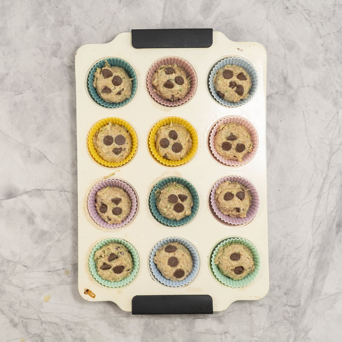Muffin tray with colourful silicone liners inside, muffin batter inside tray with 3 chocolate chips on top.