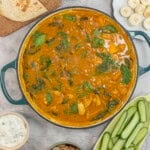 Chicken curry in pan on benchtop with sides, sliced cucumber, cashews, banana slices, naan and dip.