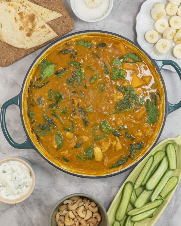 Chicken curry in pan on benchtop with sides, sliced cucumber, cashews, banana slices, naan and dip.