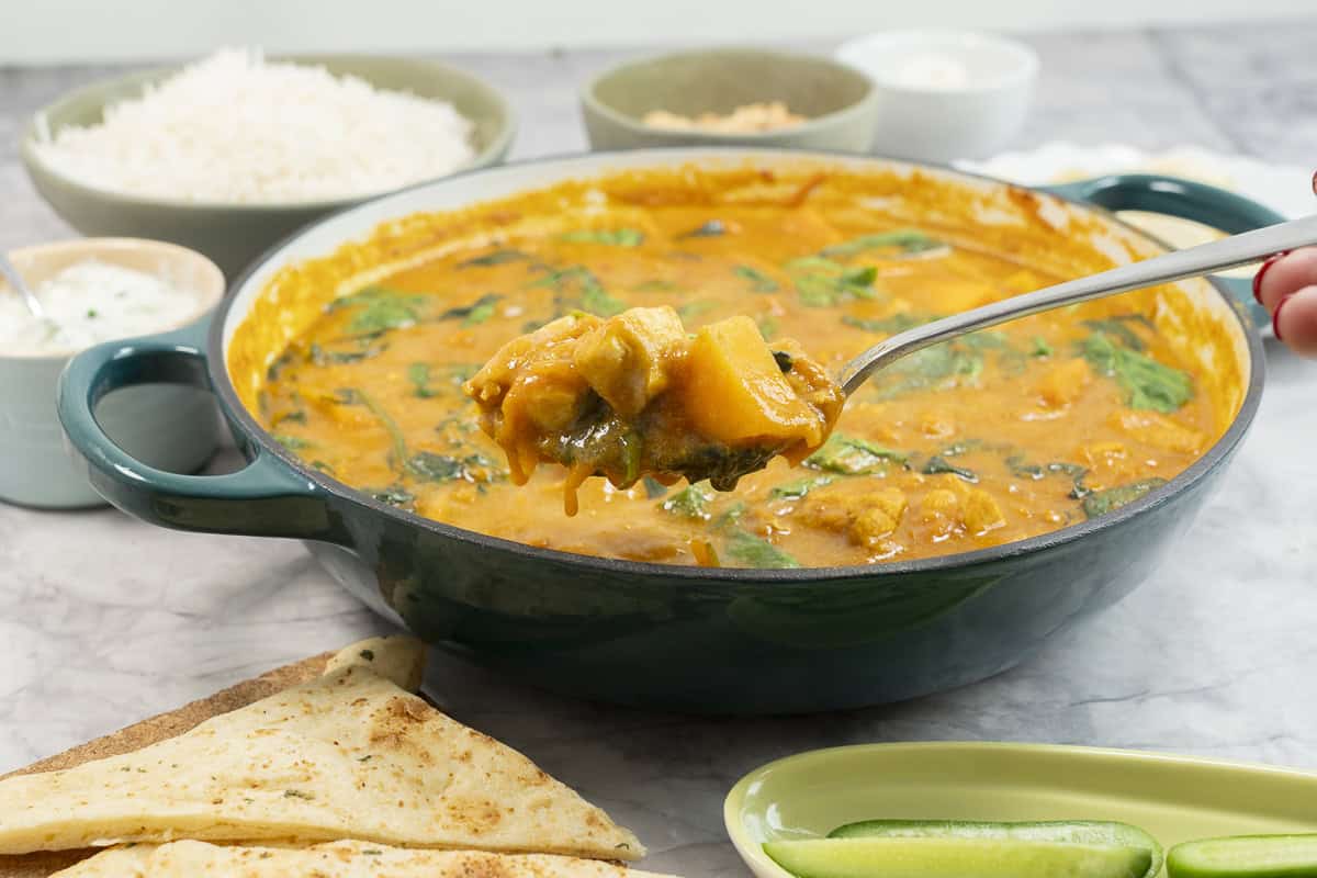 Serving spoon scooping curry out of pan on benchtop.