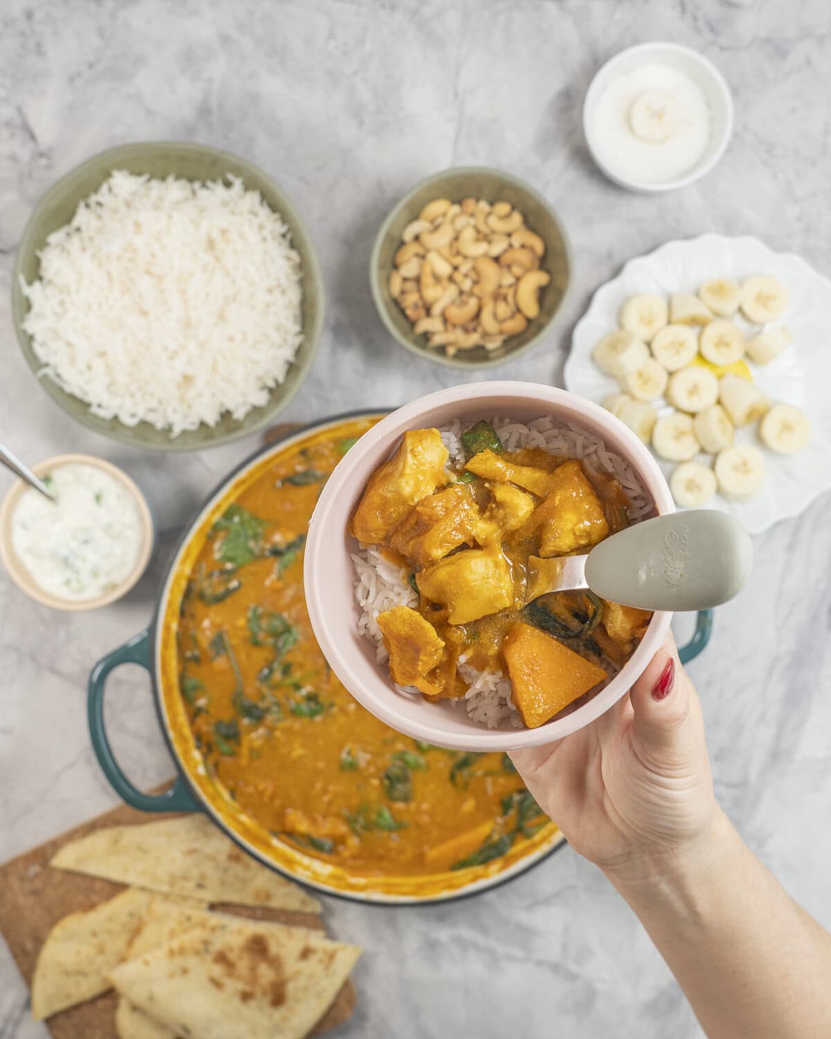 Hand holding toddler silicone plate with baby fork, rice and chicken curry inside. Sides blurred in background on benchtop.