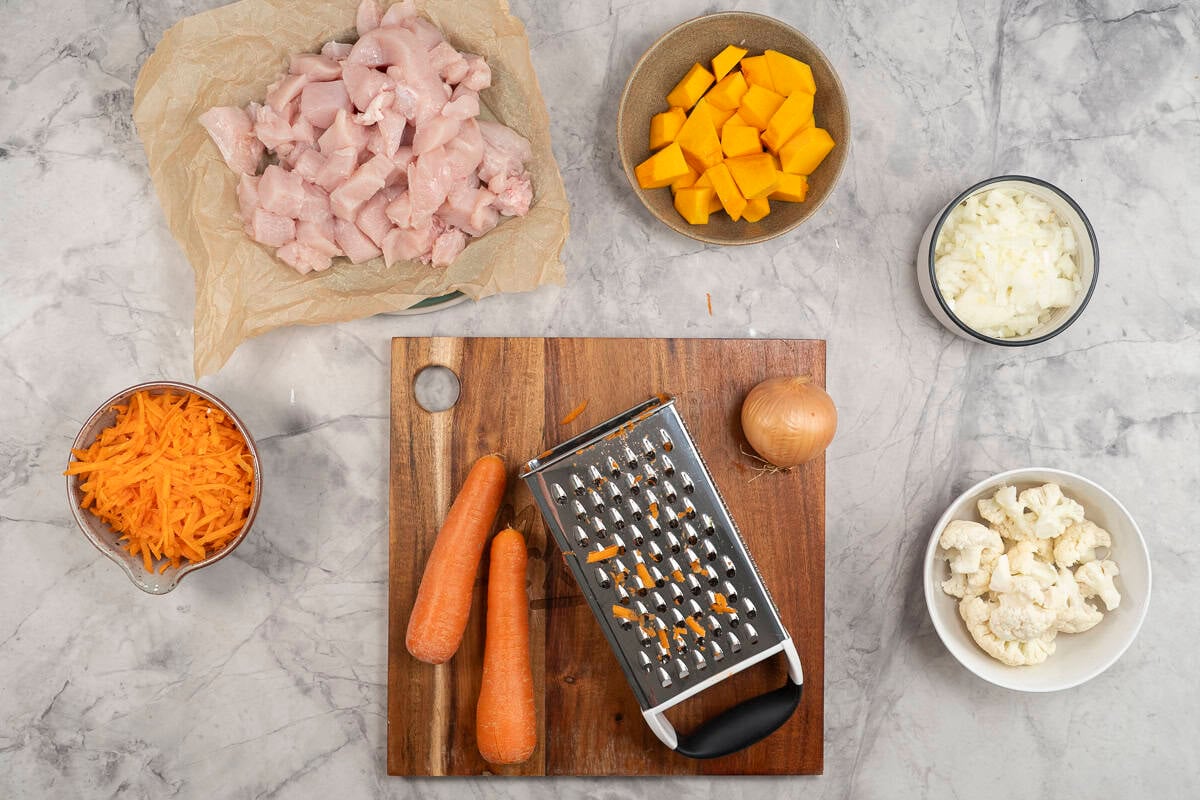 Wooden chopping board with grater, two carrots and onion. Small bowls with cauliflower chopped pumpkin, chopped chicken, diced onion and grated carrots inside.