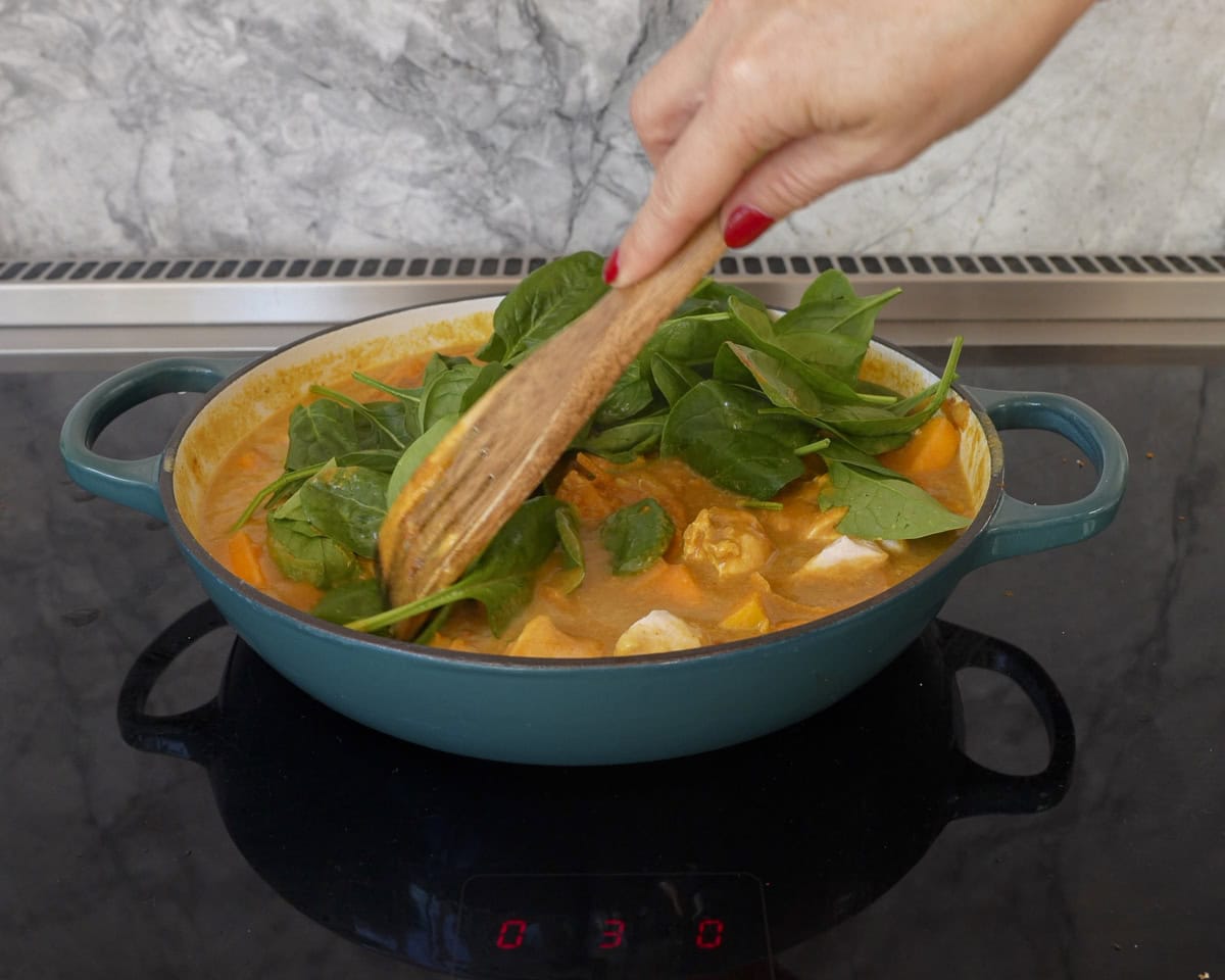 Hand holding wooden spoon mixing fresh spinach into chicken curry on stove.