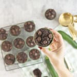 Hand holding a zucchini chocolate chip muffin in green silicone mold with more below on a cooling rack.
