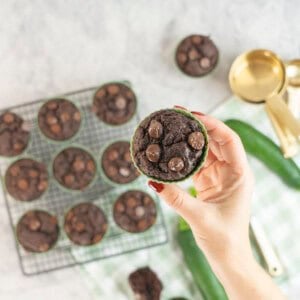 Hand holding a zucchini chocolate chip muffin in green silicone mold with more below on a cooling rack.