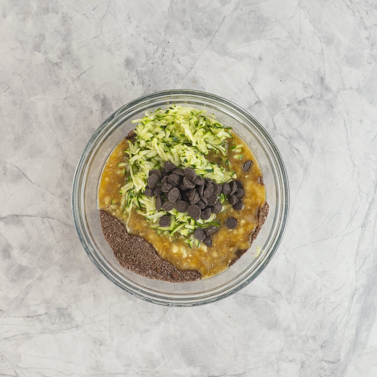 Banana mixture added into dry ingredients with grated zucchini and chocolate chips in glass bowl on benchtop.
