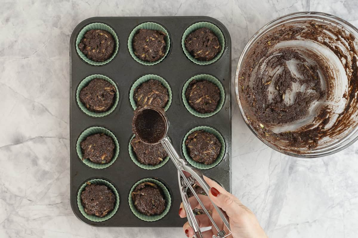 Cookie scooper scooping out mixture from glass bowl into silicone muffin cases in muffin tray.