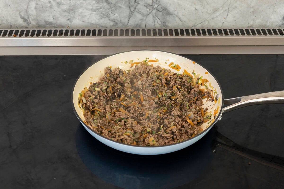 A pan cooking on stove with chopped onion, grated carrot, zucchini mushroom and mince.