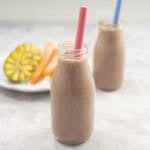 Two small glass bottles with blue and pink straw inside inside chocolate milk on benchtop. Kiwifruit and carrots on plate in background.