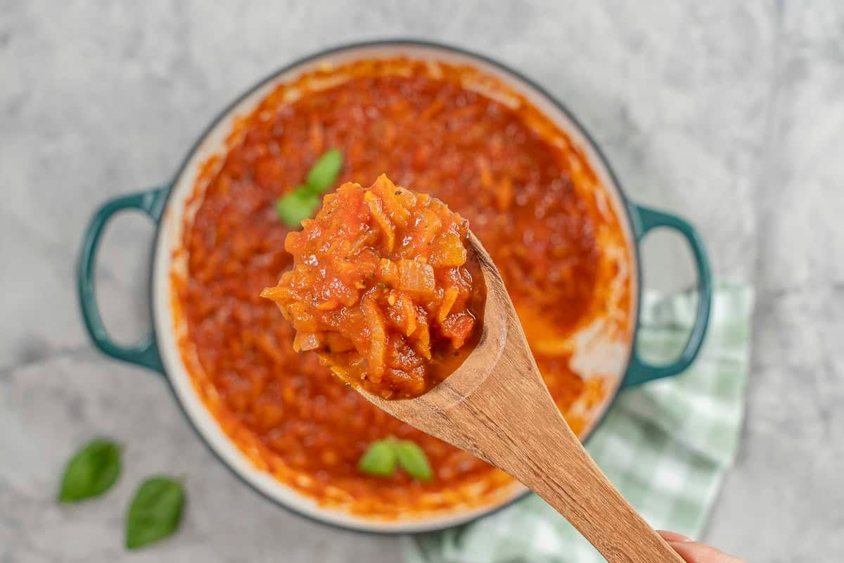 Hand holding a wooden spoon of marinara sauce over a pan of tomato sauce.