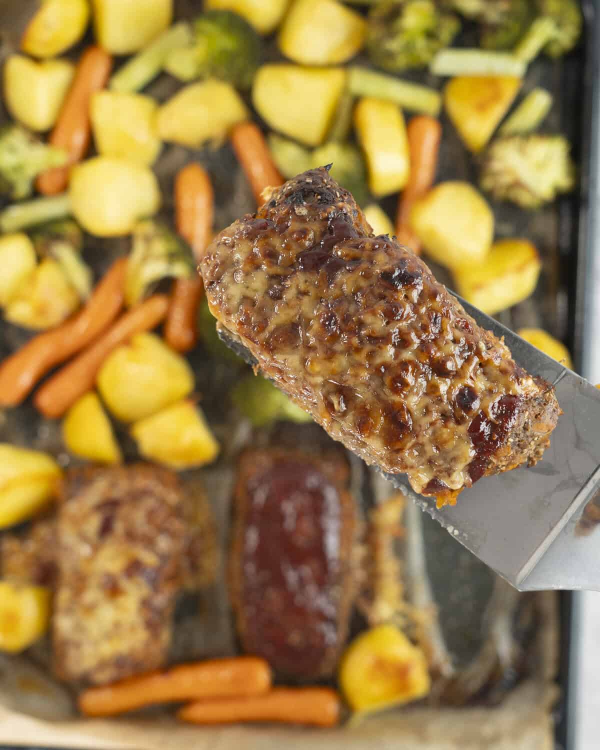 Metal spatula holding a mini meatloaf above tray of roast vegetables.