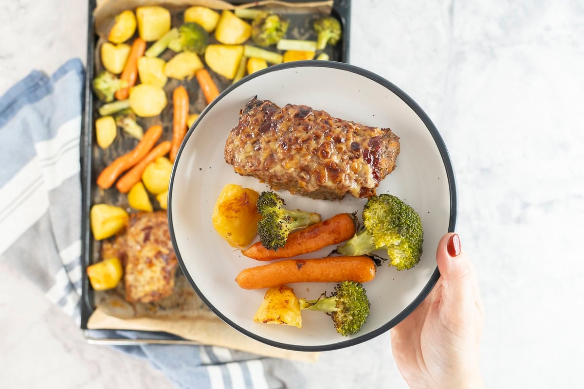 Hand holding a dinner plate with roast carrot, broccoli and potato and a small meatloaf above a lined baking tray of roast vegetables and other mini meatloaf's.