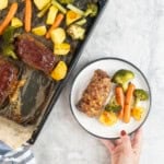 Hand holding a dinner plate with roast carrot, broccoli and potato and a small meatloaf next to a lined baking tray of roast vegetables and other mini meatloaf's.