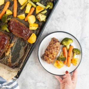 Hand holding a dinner plate with roast carrot, broccoli and potato and a small meatloaf next to a lined baking tray of roast vegetables and other mini meatloaf's.