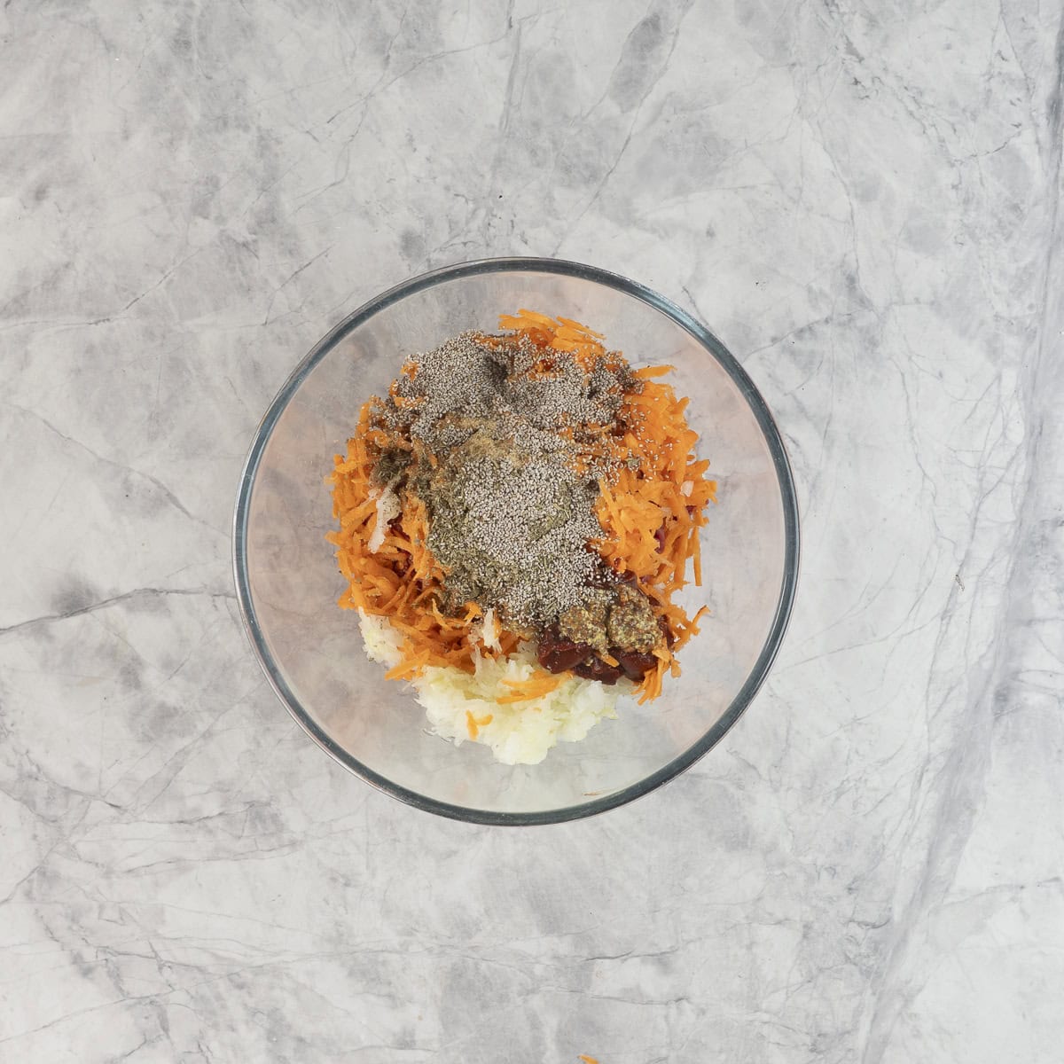Large glass bowl on benchtop with beef mince, grated carrot and onion, tomato paste, chia seeds, mustard, soy sauce, garlic powder, herbs, and ketchup.